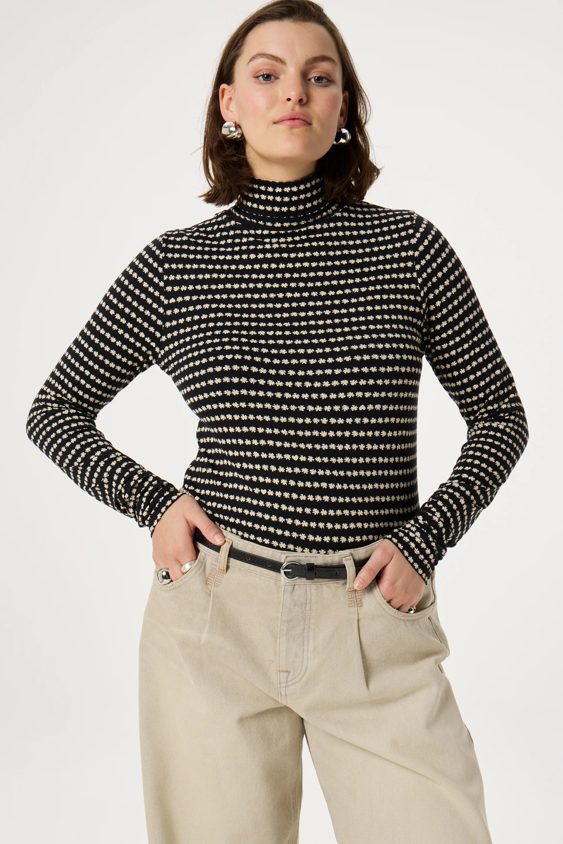 A person dressed in the Jade Top - Black by Fabienne Chapot and beige pants stands with hands in pockets against a plain background.