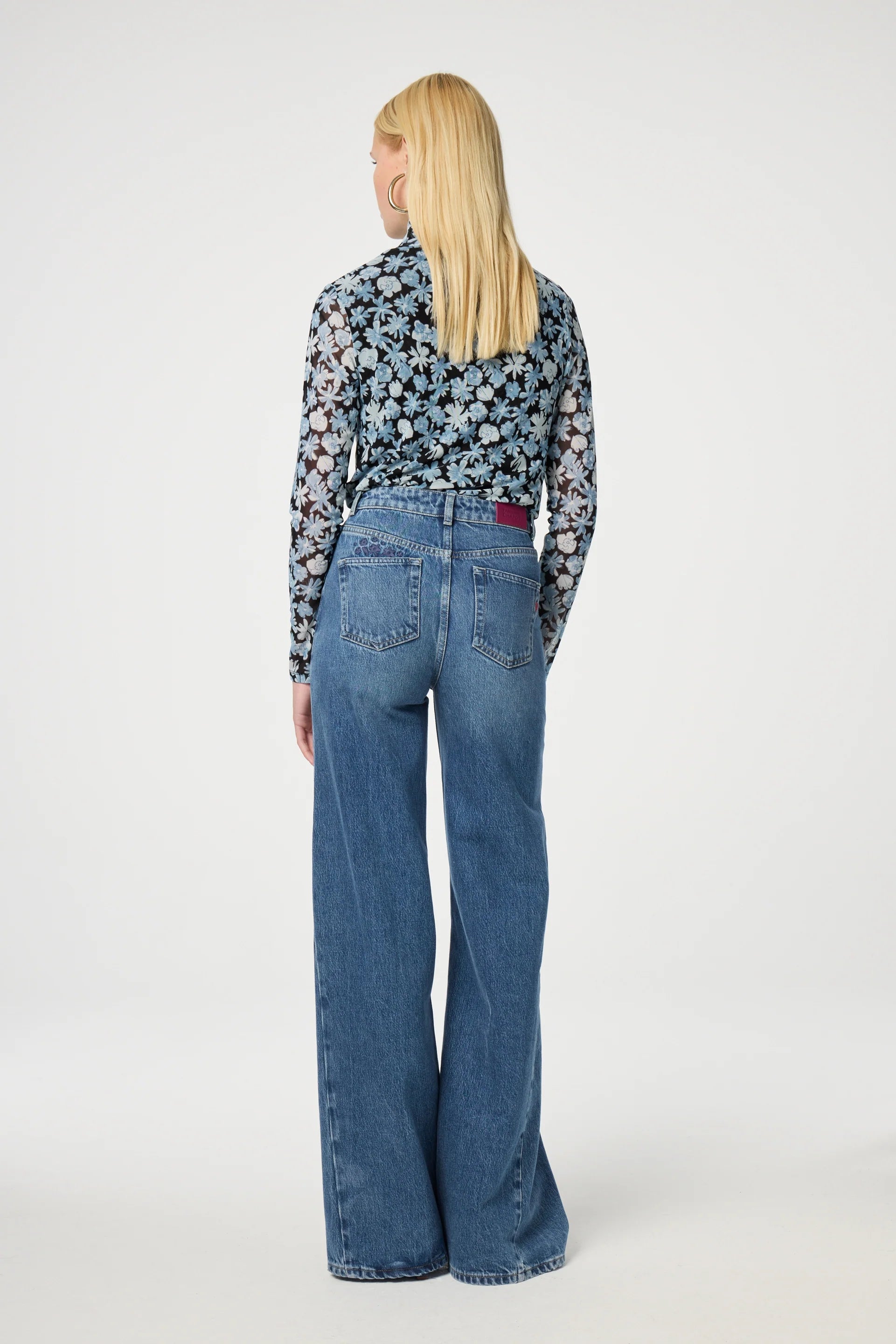 A person with long blonde hair stands with their back to the camera, wearing the Jane Top - Blue Freja from Fabienne Chapot, paired with wide-leg blue jeans in a neutral setting.