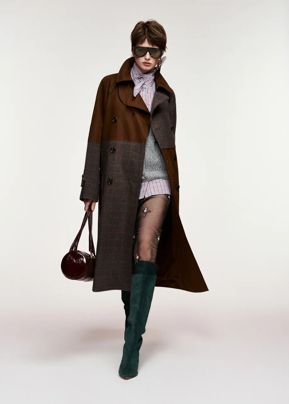 A model wearing a long brown and gray patchwork coat, a striped knit top, sheer tights, dark green knee-high boots, and carrying a dark brown leather handbag against a plain light background. Essentiel Antwerp.