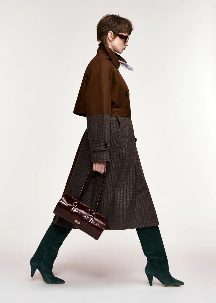 A model in side profile wearing a brown double-breasted coat over a dark brown plaid skirt, paired with dark green knee-high boots and carrying a dark brown handbag, set against a plain light gray studio background.