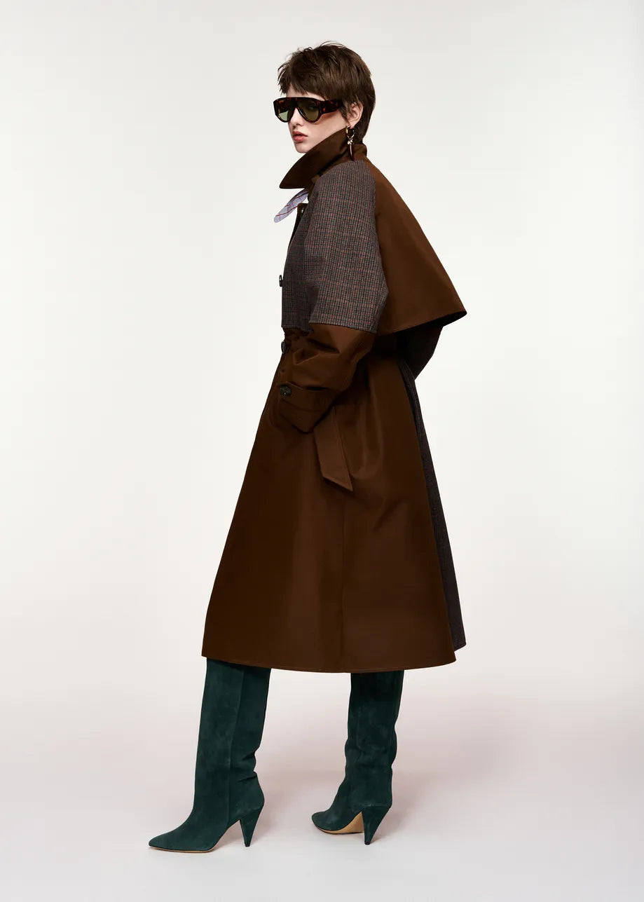 A person wearing the Essentiel Antwerp Kagu Brown and Check Trench Coat, styled with teal knee-high boots, sunglasses, and statement earrings, stands against a plain white background.