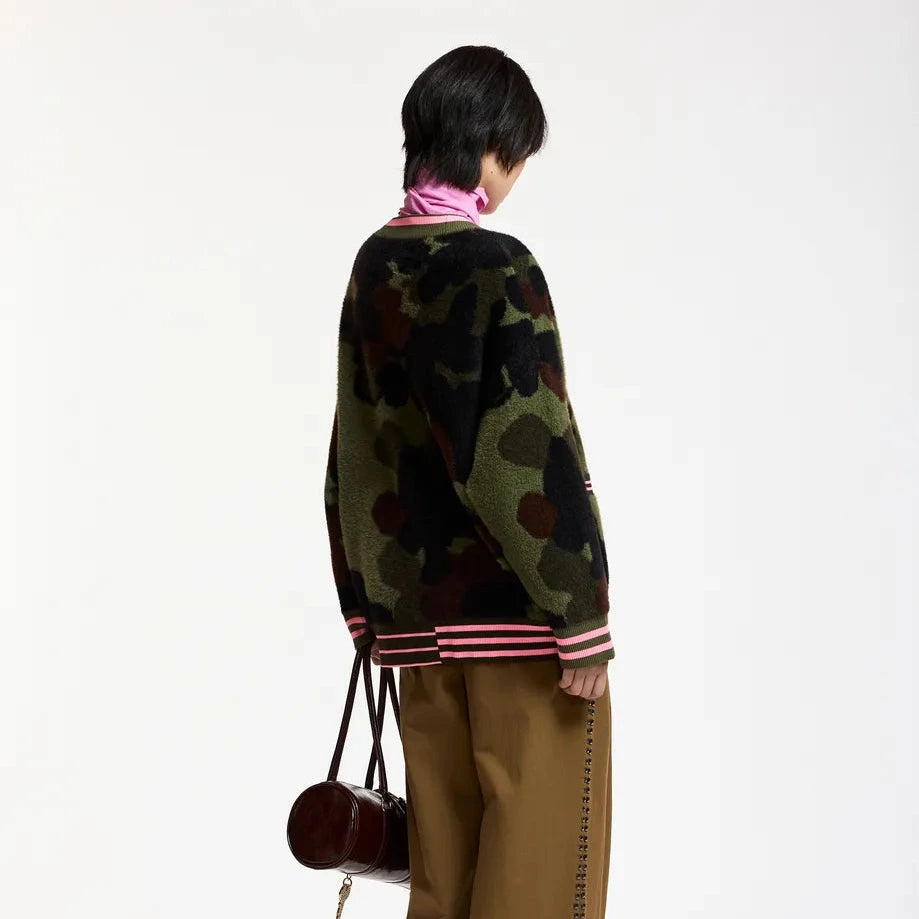 A model shown from the back wearing a green camouflage knit sweater with pink and white striped ribbed cuffs and hem, paired with wide-leg brown trousers and carrying a small dark red handbag, against a plain white studio background.