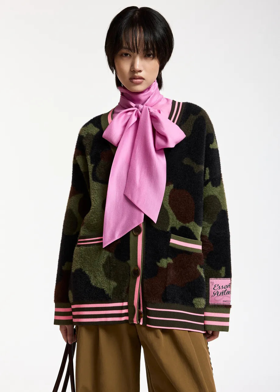 A model wearing a green and brown camouflage knit cardigan with pink striped trim, a large pink satin neck bow, and tan trousers, photographed against a plain white studio background.