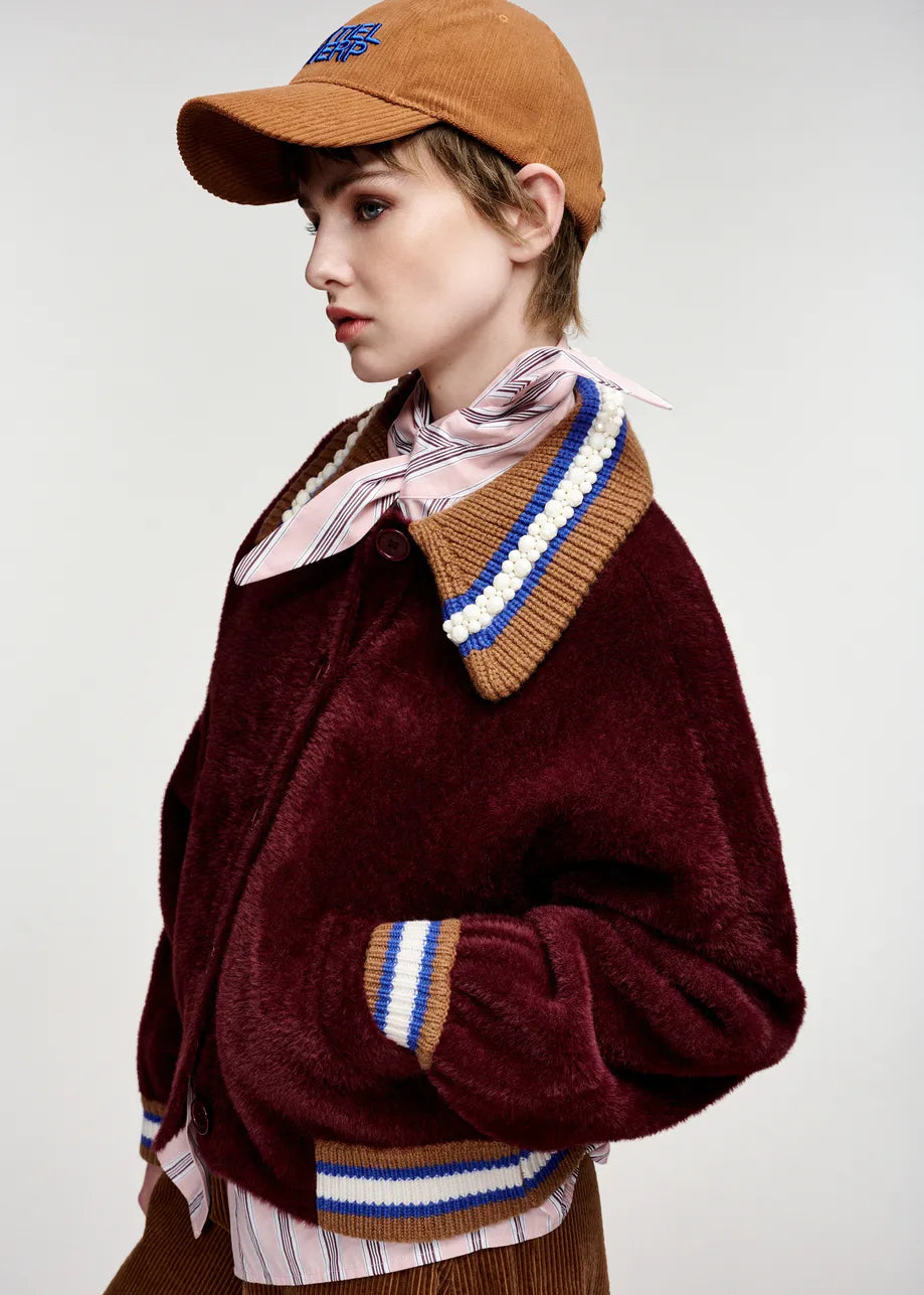 A model wearing a maroon plush fleece jacket with a striped knit collar and cuffs, a pink striped shirt, a pink neck scarf, and a brown baseball cap, photographed against a light gray studio background.