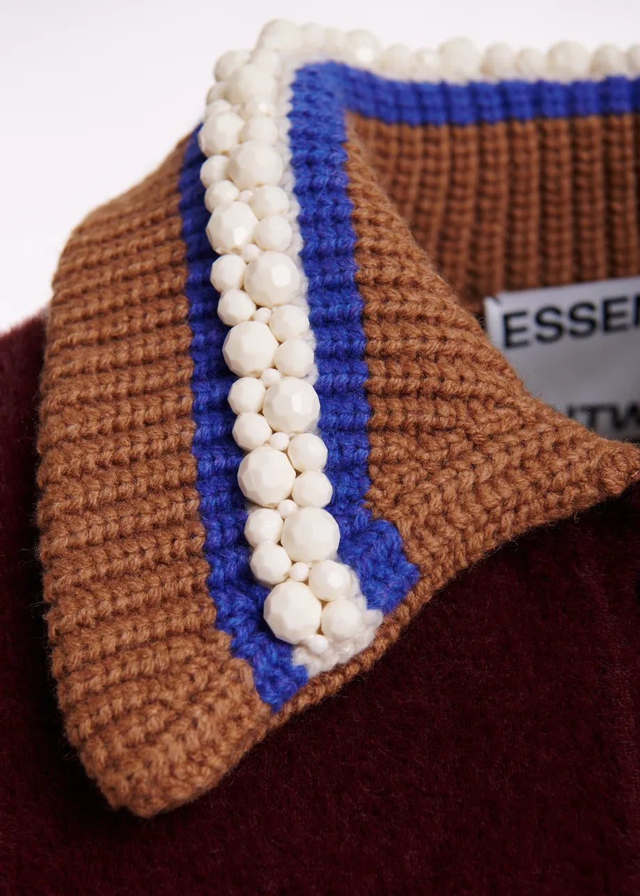 Close-up of a chunky knit sweater collar in brown and blue with a vertical row of white faux-pearl beads, resting on a burgundy fabric surface.