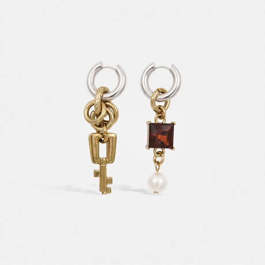 A pair of gold and silver hoop earrings with dangling charms, one featuring a gold key and the other a square dark red gemstone and a small white pearl, set against a plain white background.