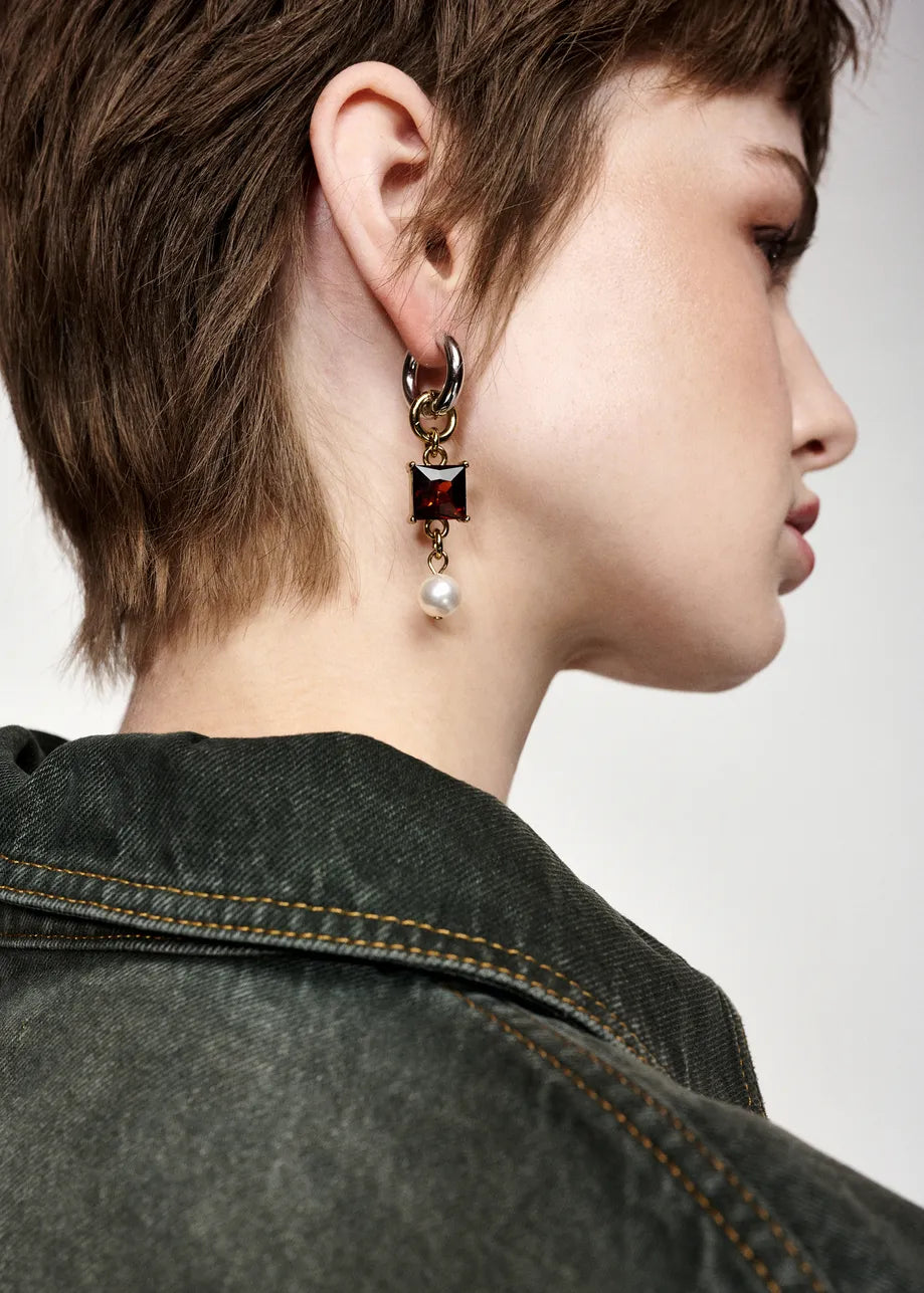 A close-up side profile of a model with short brown hair wearing a dangling earring. The earring features a gold-toned huggie hoop, a gold chain, a square dark red gemstone, and a dangling white pearl.
