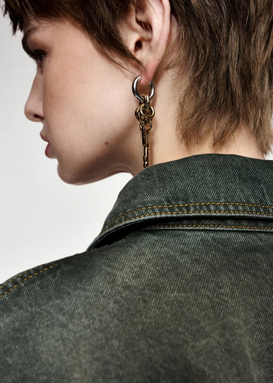 A close-up side profile of a model wearing a large, ornate gold-toned hoop earring with a dangling key charm, paired with a dark denim jacket against a light gray background.