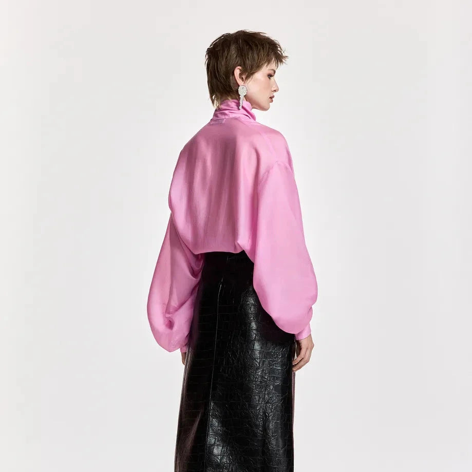 A model is shown from the back wearing a vibrant pink, voluminous long-sleeve blouse with a high neck, paired with a black faux-leather skirt and black boots, against a plain white studio background.