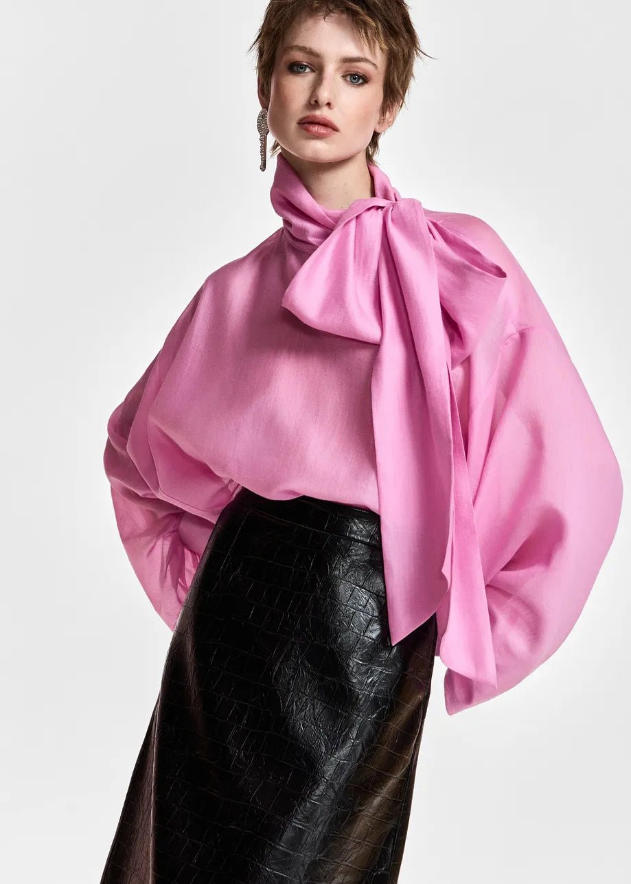 A model wearing a vibrant pink oversized blouse with a large bow tie-neck detail and voluminous balloon sleeves, paired with a black textured skirt, against a plain light gray background.