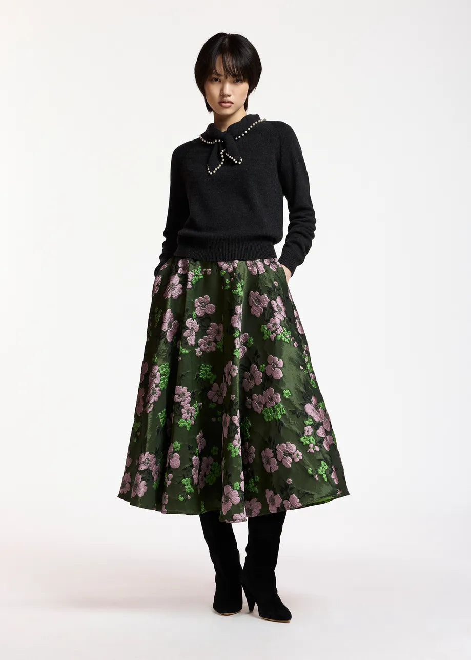 Wearing the Essentiel Antwerp Kasa Pullover with Beads in black—a wool-cashmere sweater featuring a statement collar embellished with faux pearls—paired with a green floral midi skirt and black boots, the person stands against a plain white background.