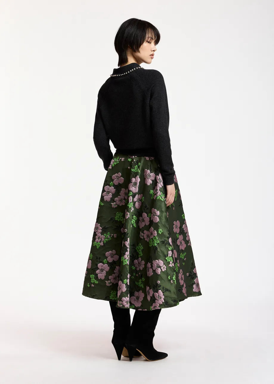A person stands with their back to the camera, wearing the Essentiel Antwerp Kasa Pullover with Beads in black—a wool-cashmere sweater with a statement collar—paired with a green floral skirt adorned with pink flowers and black boots, set against a plain white background.