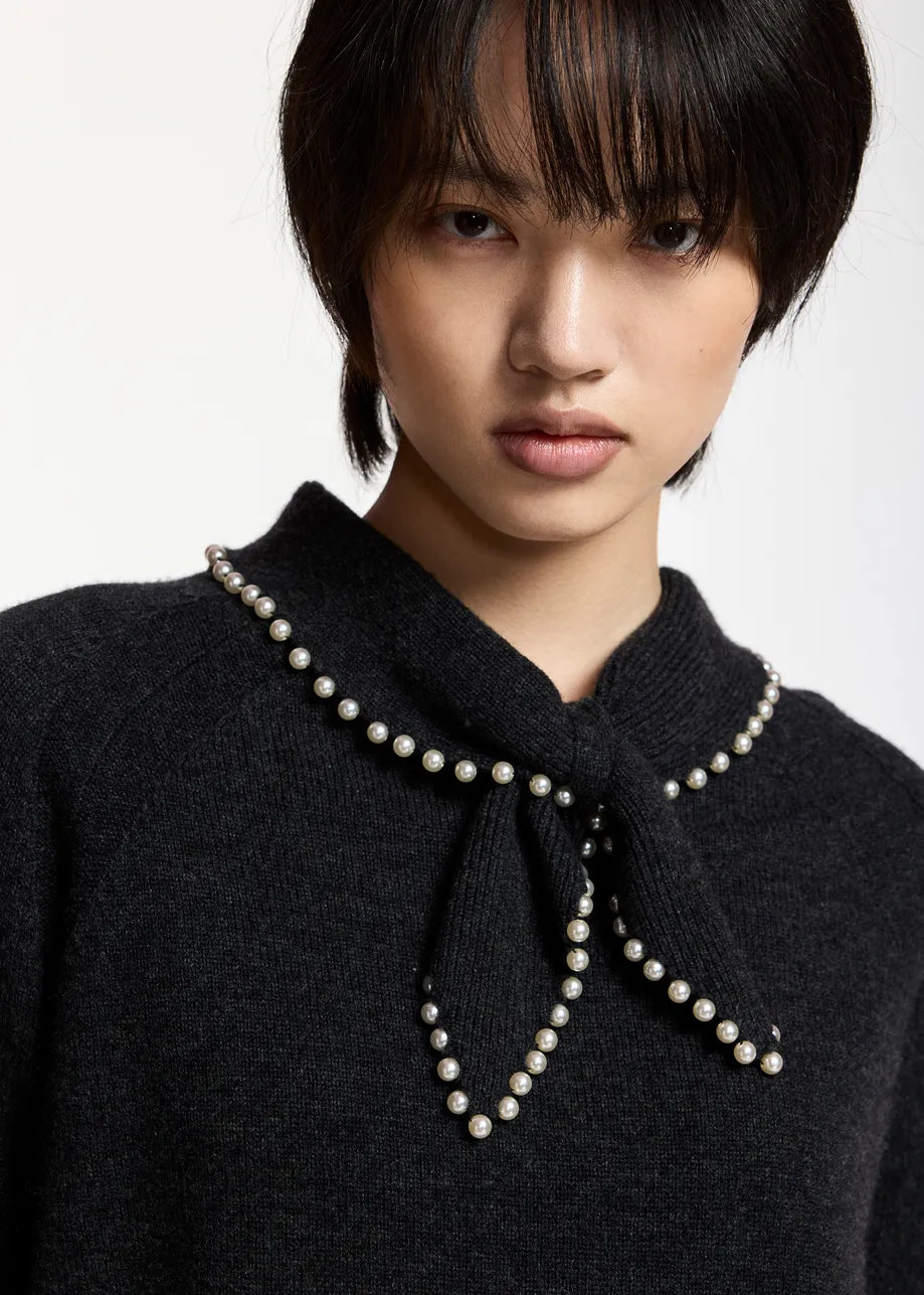 A person with short, dark hair wears the Essentiel Antwerp Kasa Pullover with Beads in black—a wool-cashmere sweater featuring a statement collar trimmed with faux pearl embellishments.