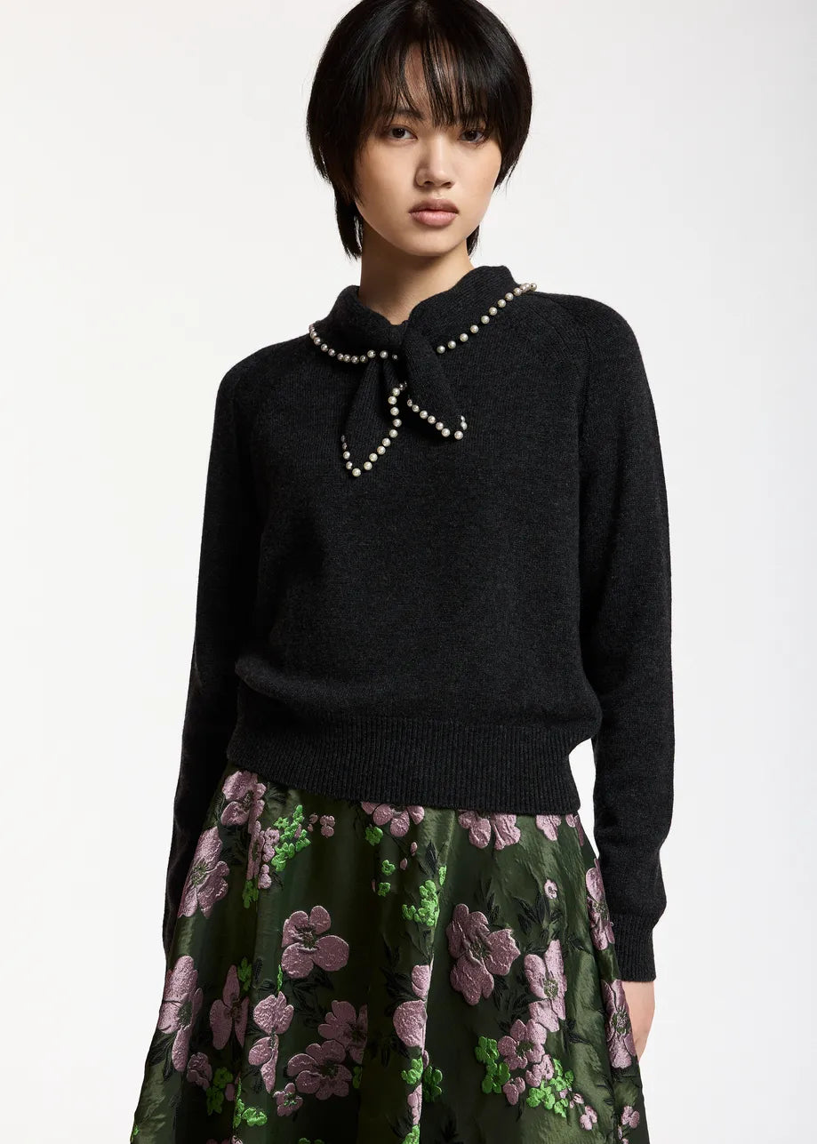 A person wearing the Essentiel Antwerp Kasa Pullover with Beads in black, featuring a statement collar embellished with faux pearls, and a green skirt with pink floral patterns stands against a plain background.
