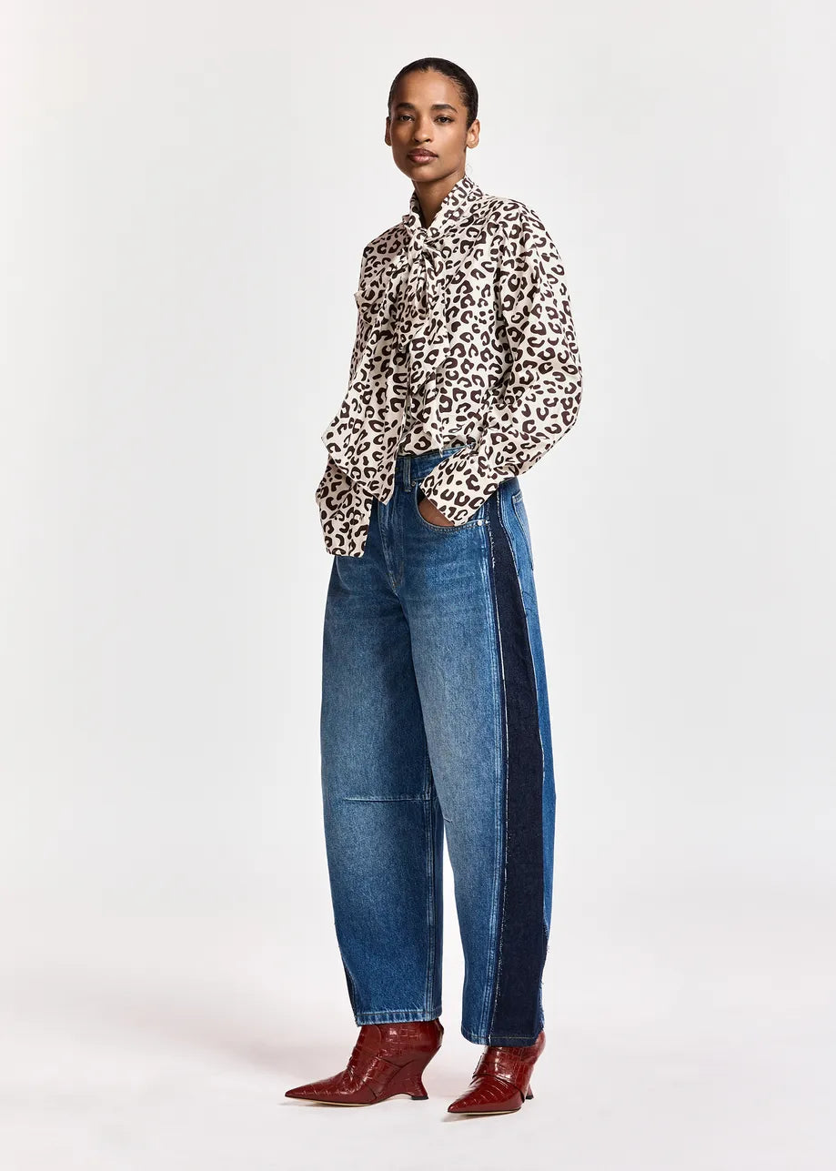 A model stands against a light gray studio background wearing a leopard-print button-up shirt, barrel-leg blue jeans with dark side panels, and brown pointed-toe boots.