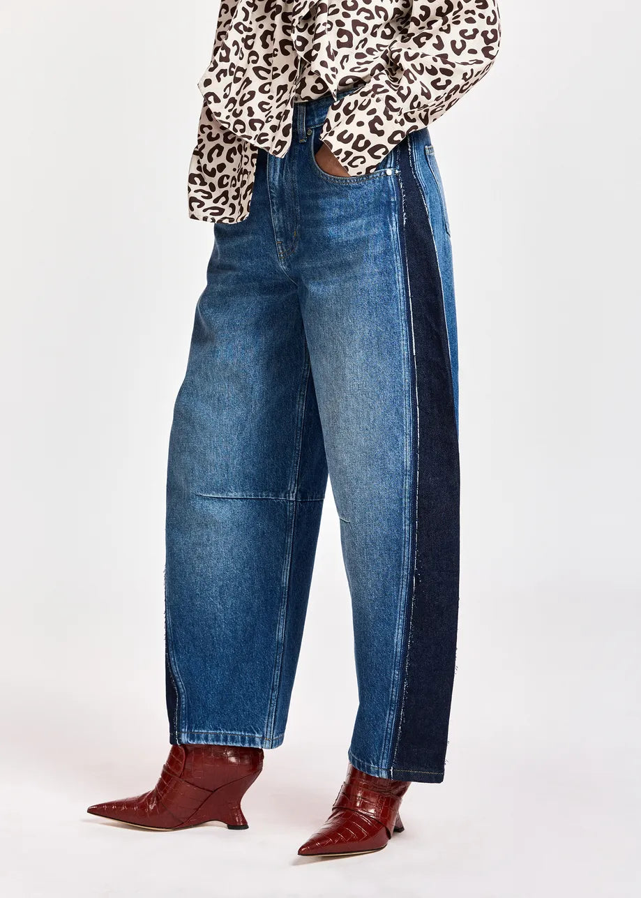 A person wearing barrel-leg blue denim jeans with dark navy side panels, paired with a leopard-print top and red pointed-toe heeled boots, shown against a plain light gray studio background.