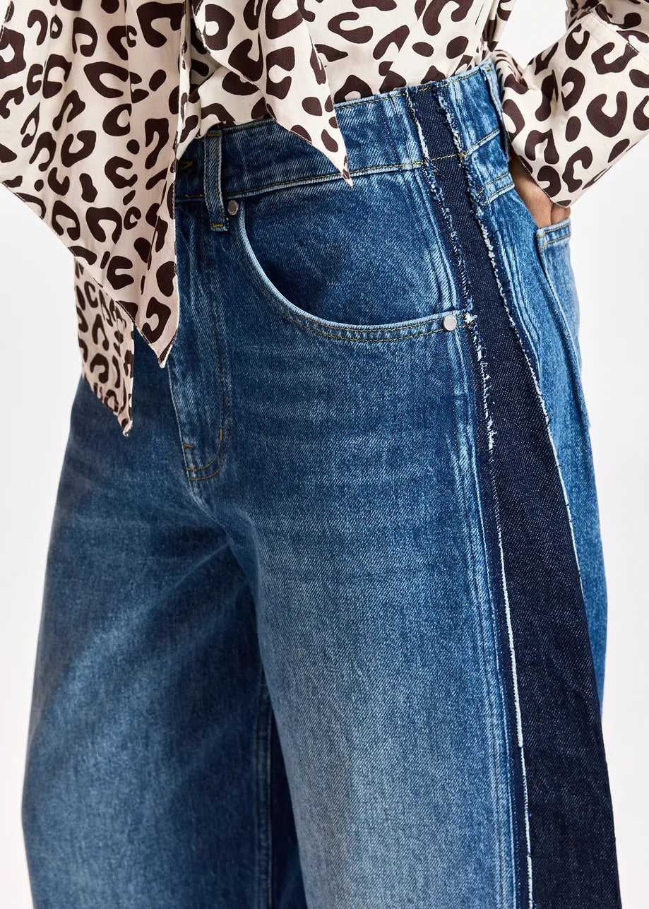 Wearing the Kashba Barrel Jeans with Panel by Essentiel Antwerp, featuring high-waisted blue denim with a dark side stripe and raw-edge detailing, paired with a cream blouse adorned in a brown animal print.