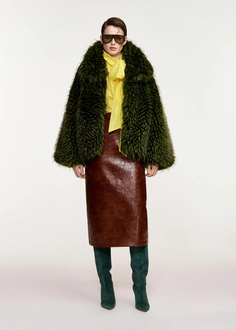 A person wearing dark sunglasses, a yellow blouse with a bow, a brown leather skirt, teal knee-high boots, and the Kastanje Oversized Faux Fur Jacket in green by Essentiel Antwerp, standing against a plain white background.