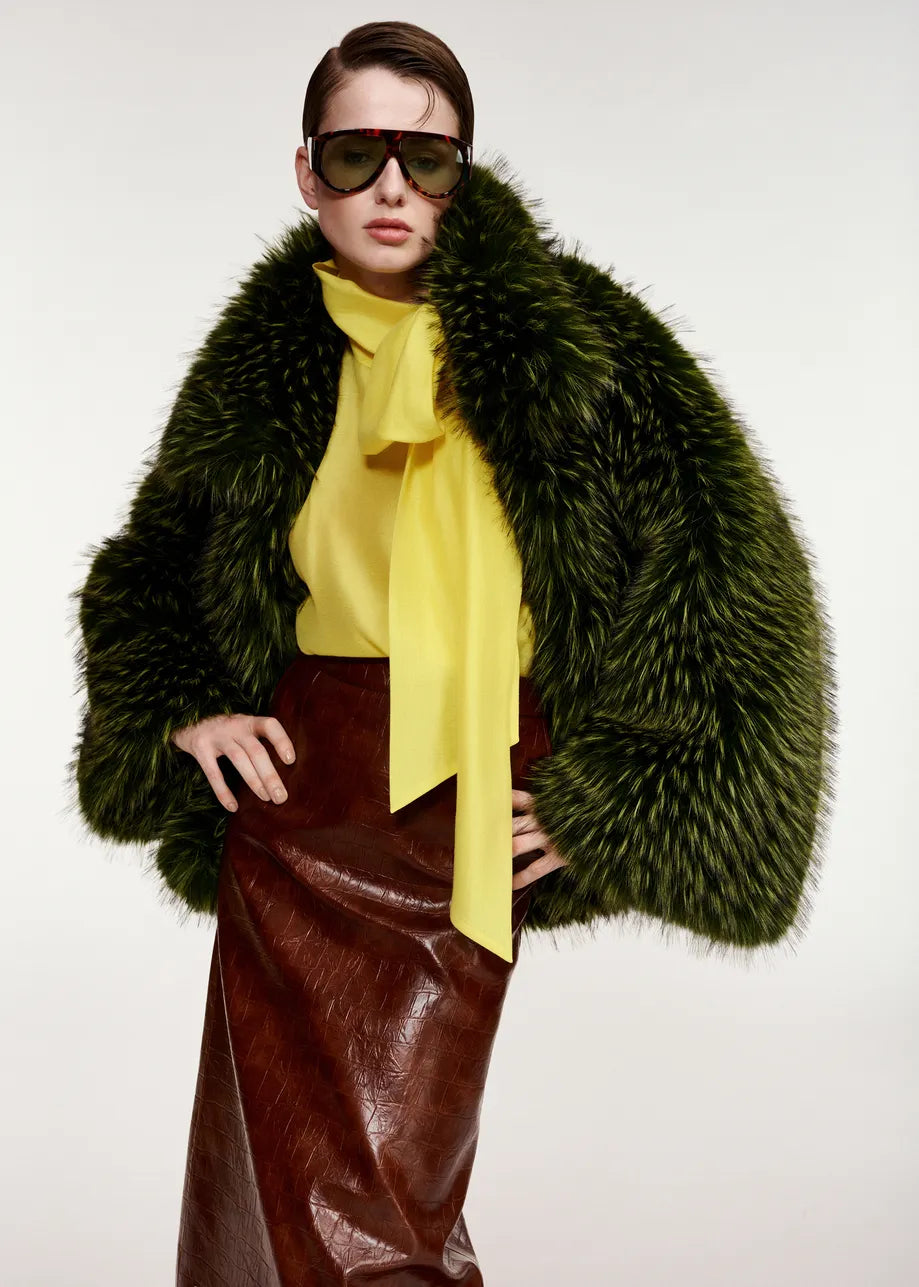 Wearing the Kastanje Oversized Faux Fur Jacket in green by Essentiel Antwerp, paired with a chunky-collared design, a yellow bow blouse, a brown leather skirt, and large sunglasses, this person poses against a plain background.