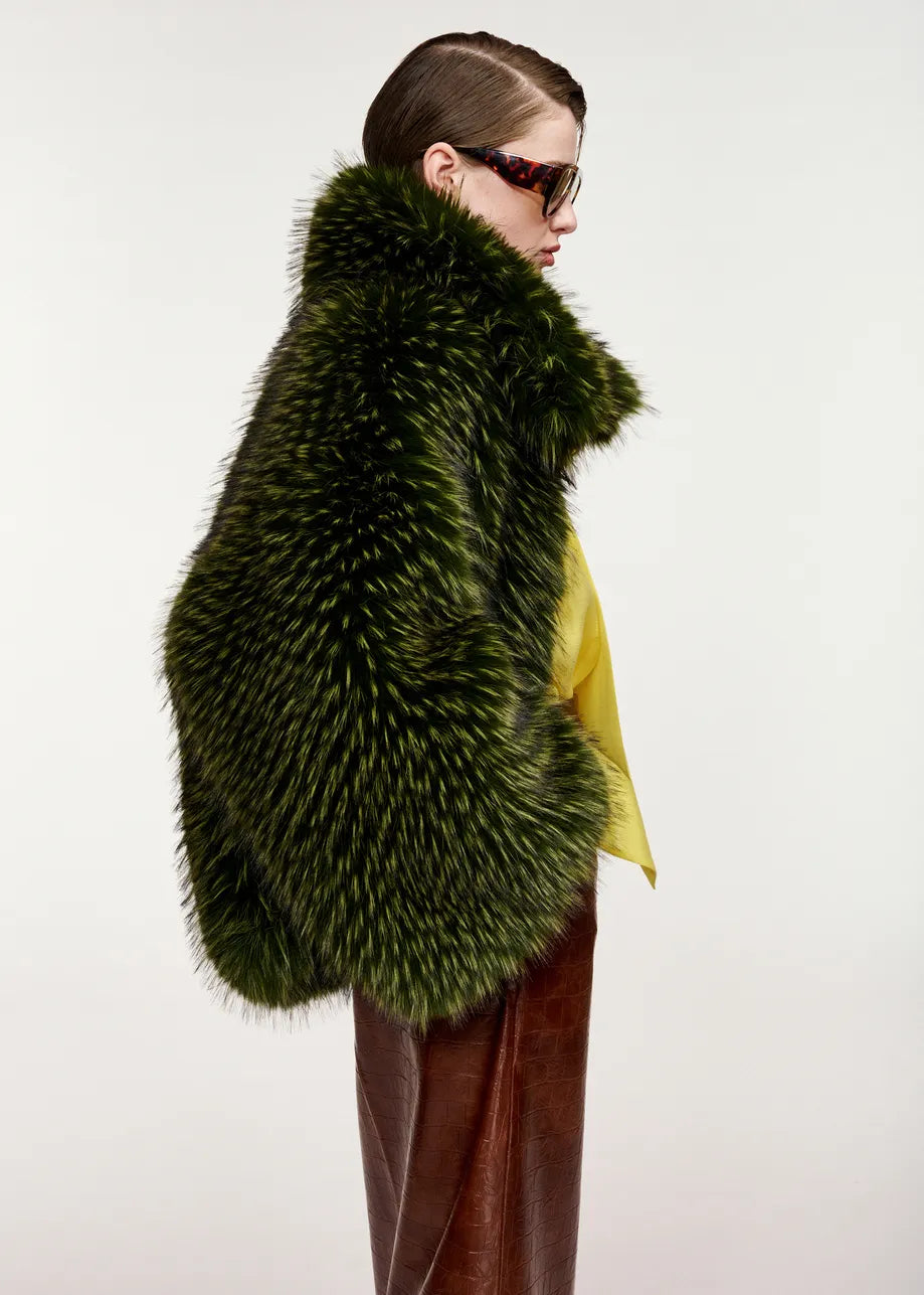 A person in side profile wears the Essentiel Antwerp Kastanje Oversized Faux Fur Jacket in green, featuring a chunky collar, styled with a yellow top, brown skirt, and sunglasses against a plain white background.