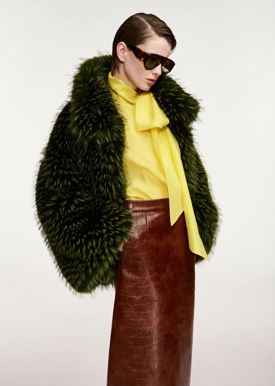 A person wearing the Kastanje Oversized Faux Fur Jacket in green by Essentiel Antwerp, featuring a chunky collar, paired with a yellow bow blouse, brown leather skirt, and large sunglasses, stands against a plain background.