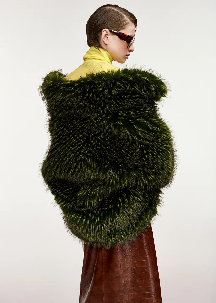 Wearing the Essentiel Antwerp Kastanje Oversized Faux Fur Jacket in green, a person in sunglasses and a yellow turtleneck pairs the statement piece with a brown skirt, posing against a plain background.
