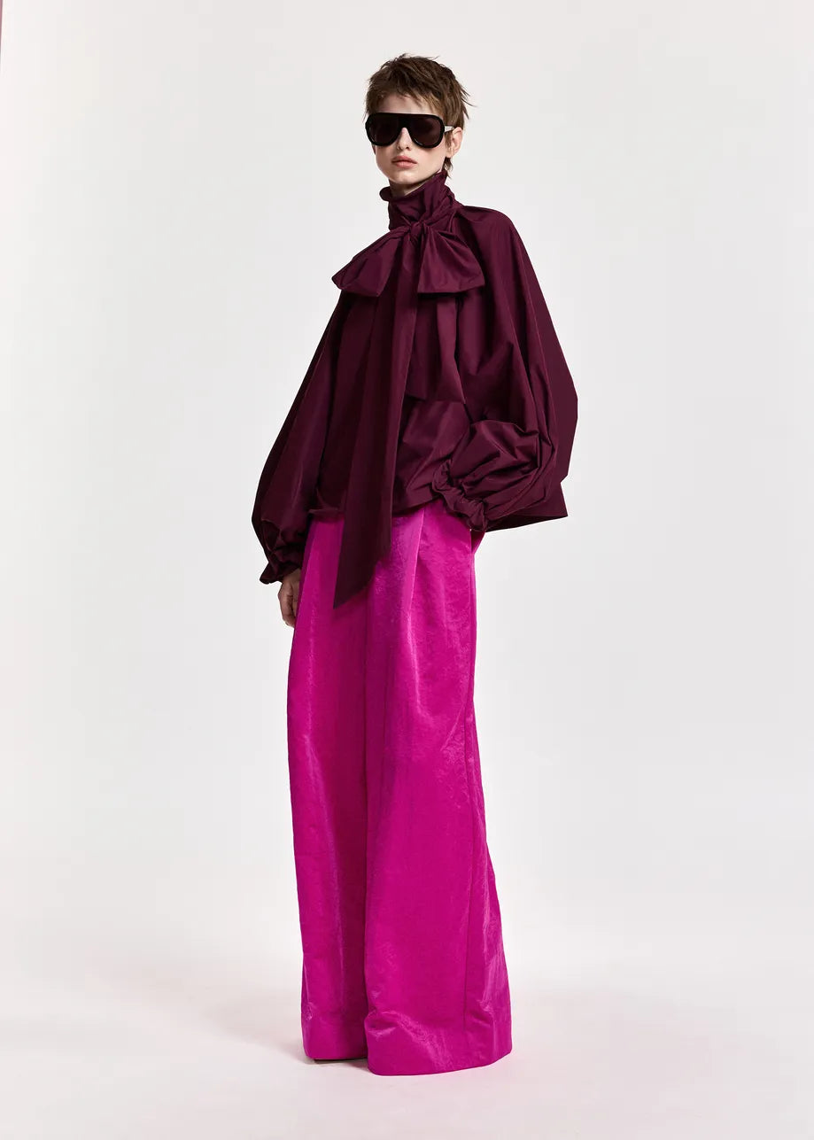 A person wearing the Essentiel Antwerp Keily Pussy Bow Tie Top in burgundy, styled with a large bow and wide magenta pants, accessorized with dark sunglasses and standing against a plain white background.