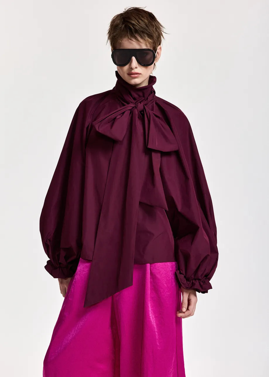 A person wearing black sunglasses, the Essentiel Antwerp Keily Pussy Bow Tie Top in Burgundy, and a shiny bright pink skirt made from recycled polyester is posed against a plain white background.