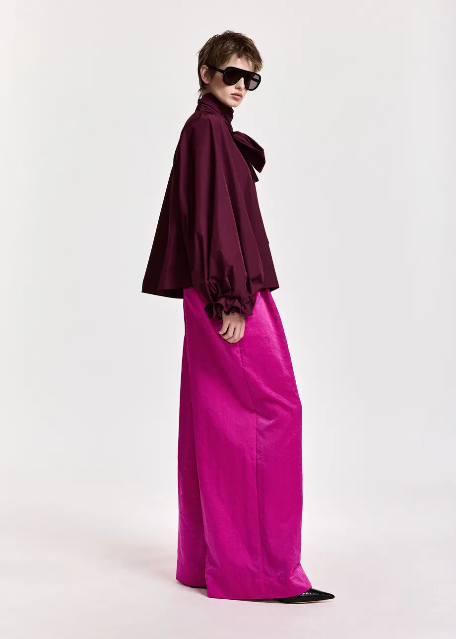 Model stands in profile against a plain light background, wearing the Essentiel Antwerp Keily Pussy Bow Tie Top in burgundy—an oversized design featuring puffy sleeves and a distinctive pussy-bow collar—paired with bright pink wide-leg pants made from recycled polyester, black pointed shoes, and dark sunglasses.