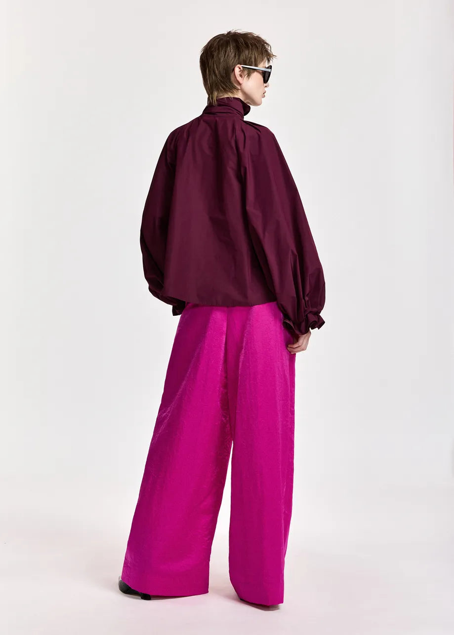 A person stands with their back to the camera, wearing the Essentiel Antwerp Keily Pussy Bow Tie Top in burgundy, paired with bright pink wide-leg pants, against a plain light background.