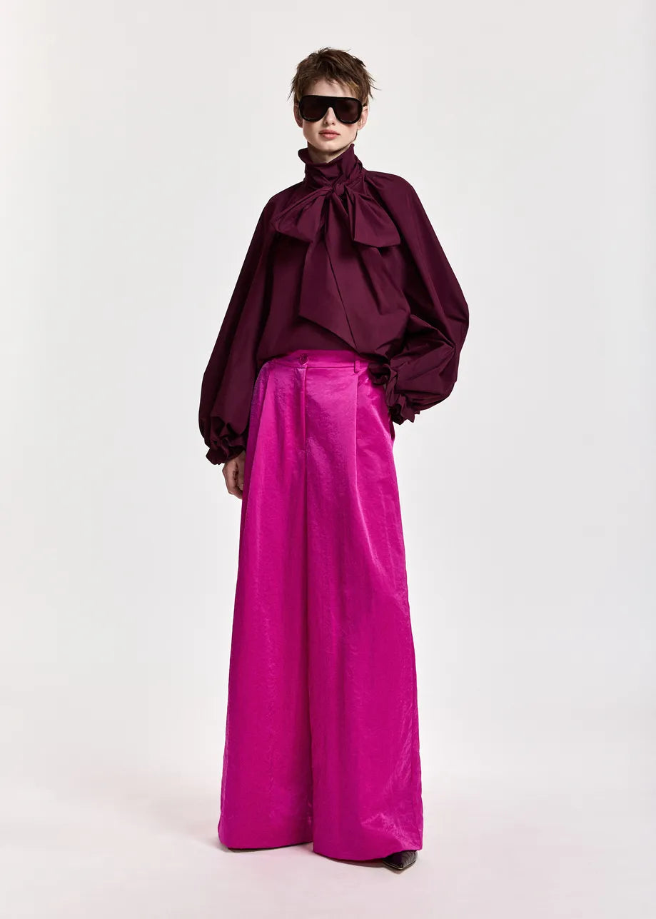 A person wearing the Keily Pussy Bow Tie Top in burgundy by Essentiel Antwerp, styled with wide-legged bright pink pants and large sunglasses, stands against a plain light background.