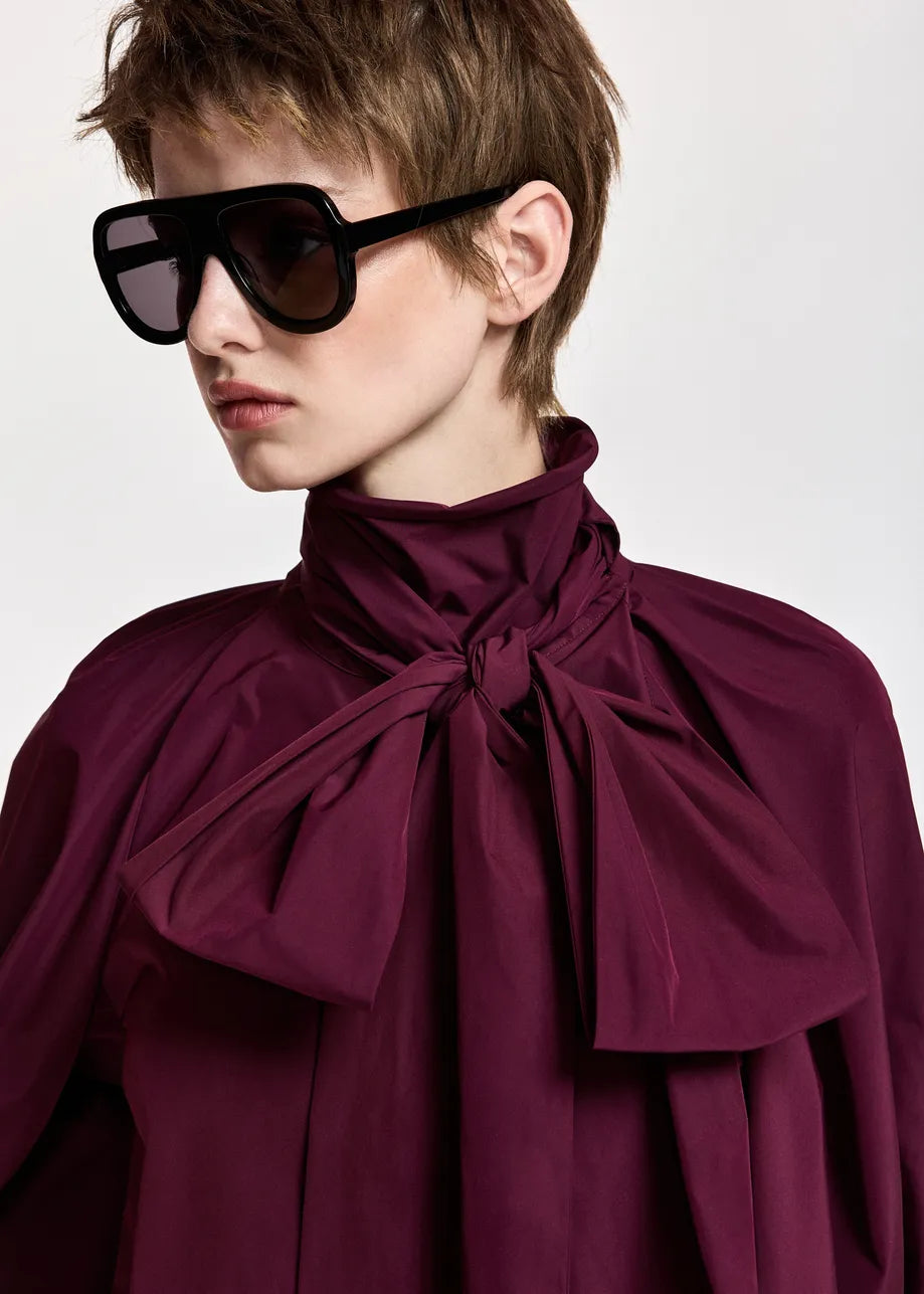 A person wearing oversized black sunglasses and the Essentiel Antwerp Keily Pussy Bow Tie Top in burgundy, featuring a high neck and large pussy-bow collar, looks to the side against a plain background.