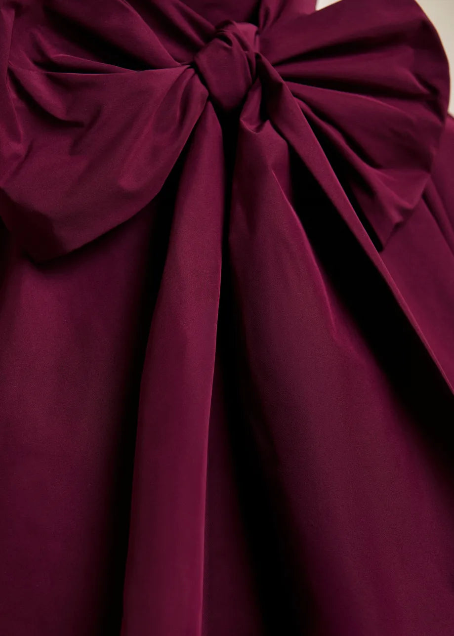 Close-up of the Keily Pussy Bow Tie Top in Burgundy by Essentiel Antwerp, featuring a large, deep burgundy recycled polyester bow with draped tails—evoking the elegance of an oversized pussy-bow collar—set against a matching background.