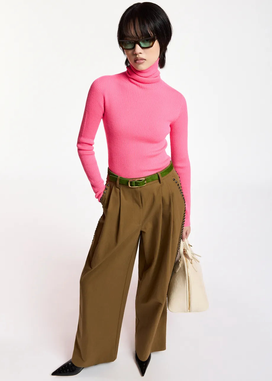 Wearing the Essentiel Antwerp Kelmuse Knitted Pullover in pink, this person pairs it with wide-leg brown trousers and a green belt, black pointed shoes, green sunglasses, and carries a beige bag while standing against a white background.