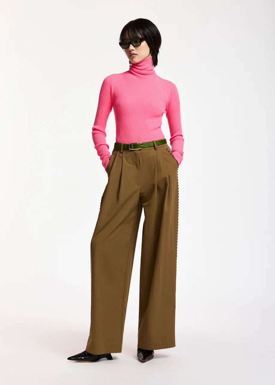 A person wearing the Essentiel Antwerp Kelmuse Knitted Pullover in pink, paired with wide-legged brown pants, a green belt, black pointed shoes, and sunglasses, stands against a plain white background.