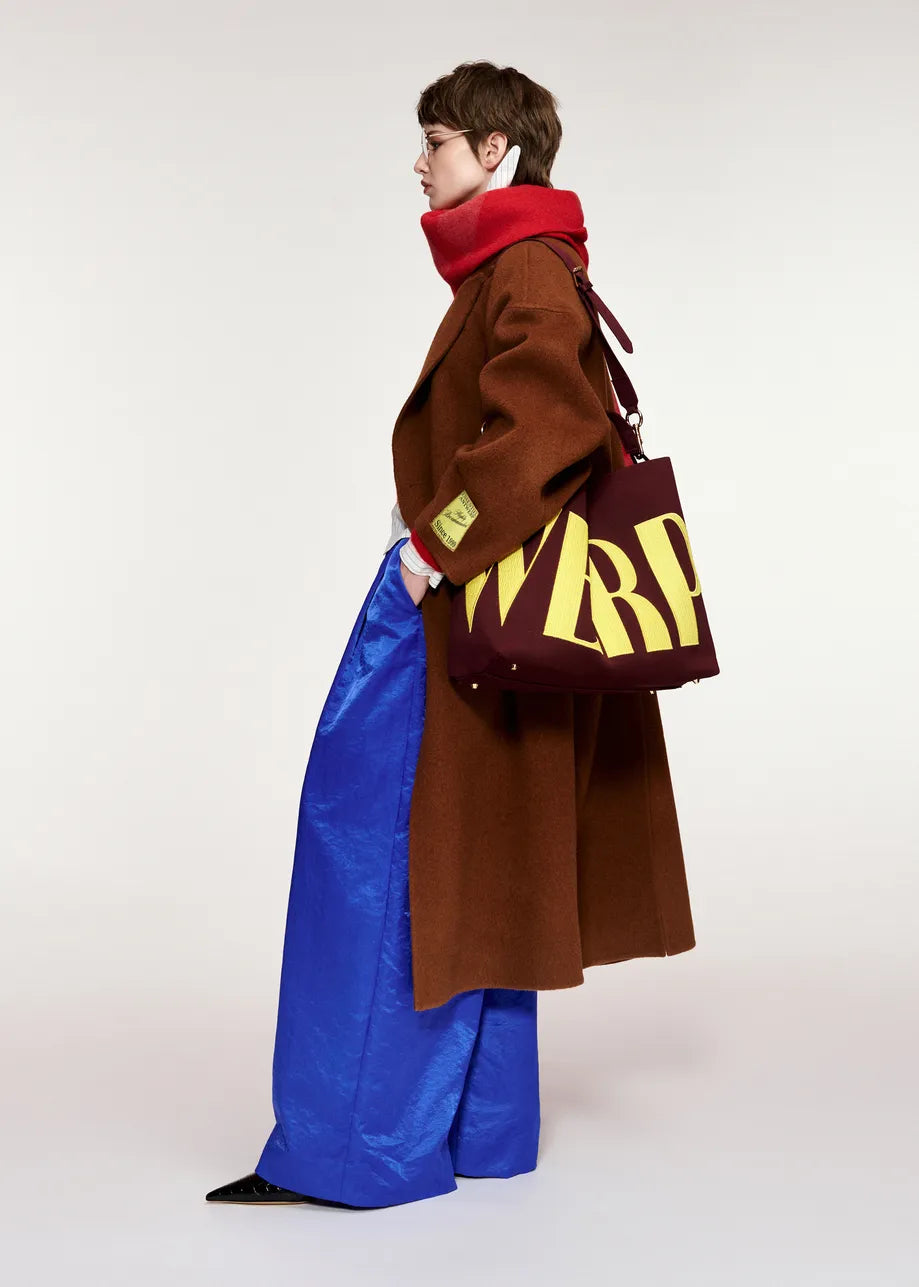 Person in the Essentiel Antwerp Kelp Oversized Wool-Cashmere Blend Coat in brown, blue wide-leg pants, and a red scarf stands in profile, holding a large burgundy tote bag with bold yellow “WRP” lettering.