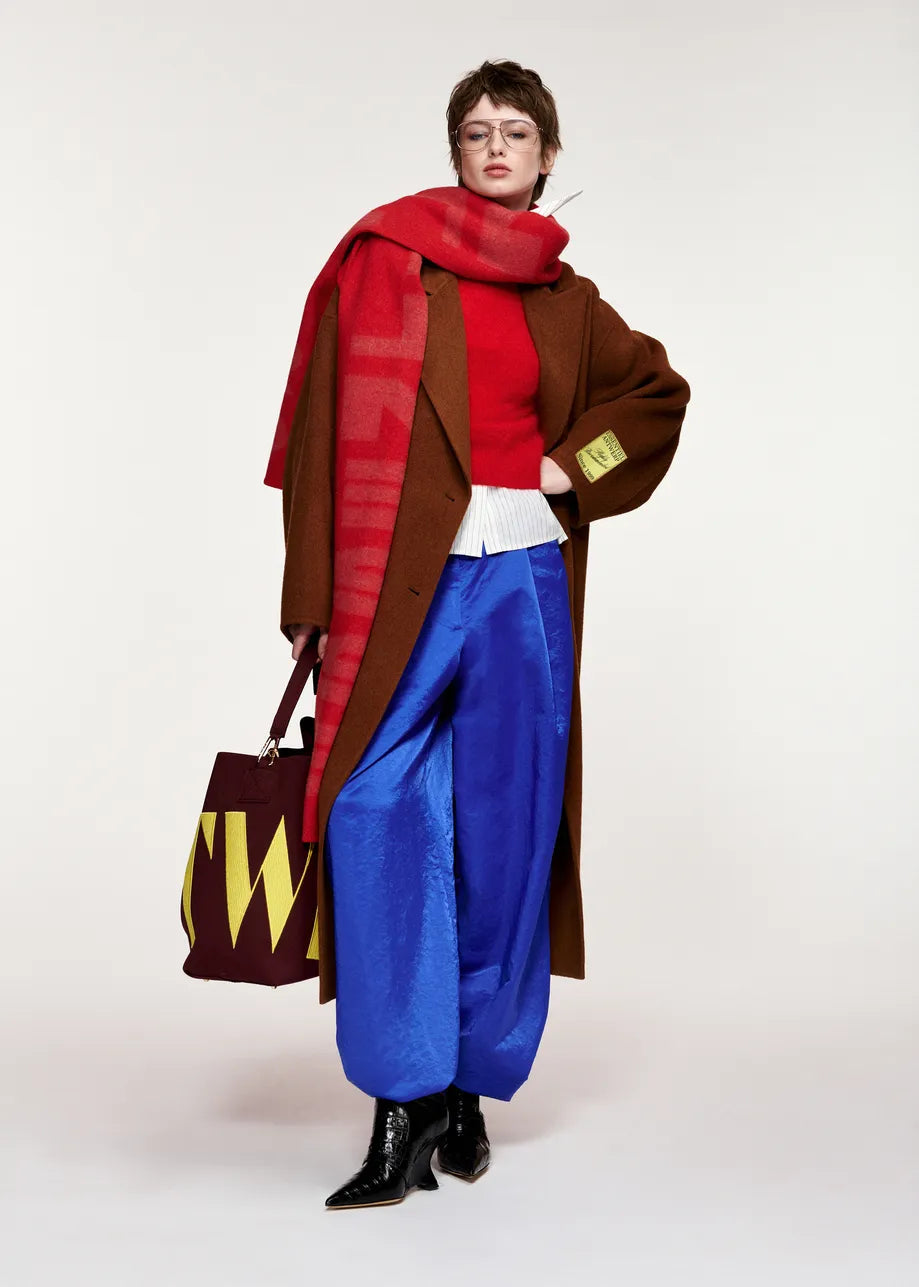 A person wearing the Essentiel Antwerp Kelp Oversized Wool-Cashmere Blend Coat in brown, paired with a red sweater, large red scarf, blue wide-leg pants, black boots, and holding a large tote bag with yellow letters.