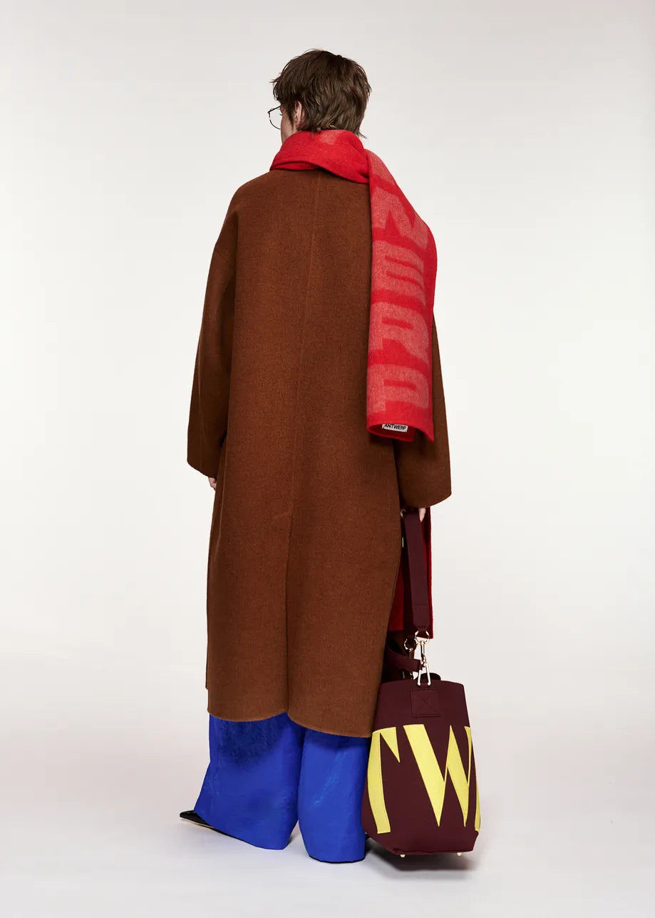 A person seen from behind wearing the Essentiel Antwerp Kelp Oversized Wool-Cashmere Blend Coat in brown, styled with a red scarf, blue pants, and holding a large burgundy bag with bold yellow letters against a plain light background.