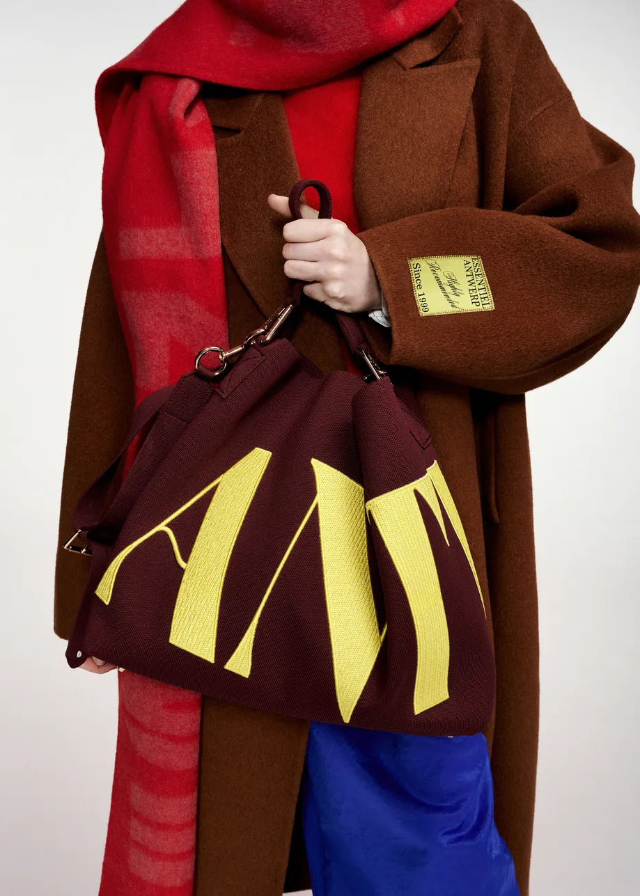 A person wearing the Essentiel Antwerp Kelp Oversized Wool-Cashmere Blend Coat in brown, styled with a red scarf and blue pants, holds a maroon bag with large yellow letters. A yellow label is attached to the coat sleeve.