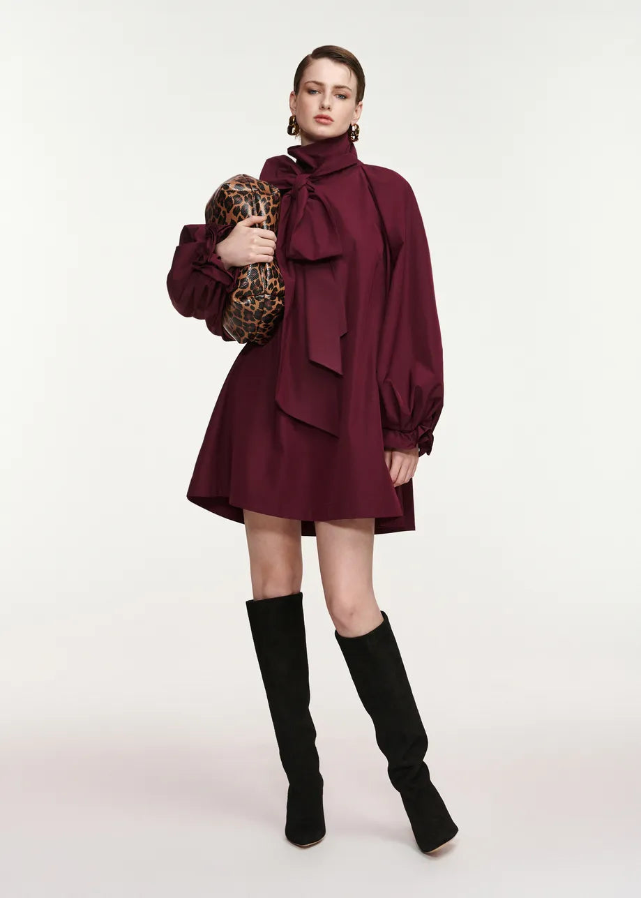 A model stands against a light gray studio background wearing a burgundy high-neck mini dress with a large bow at the collar, voluminous long sleeves, and black over-the-knee boots. She is holding a leopard-print clutch.