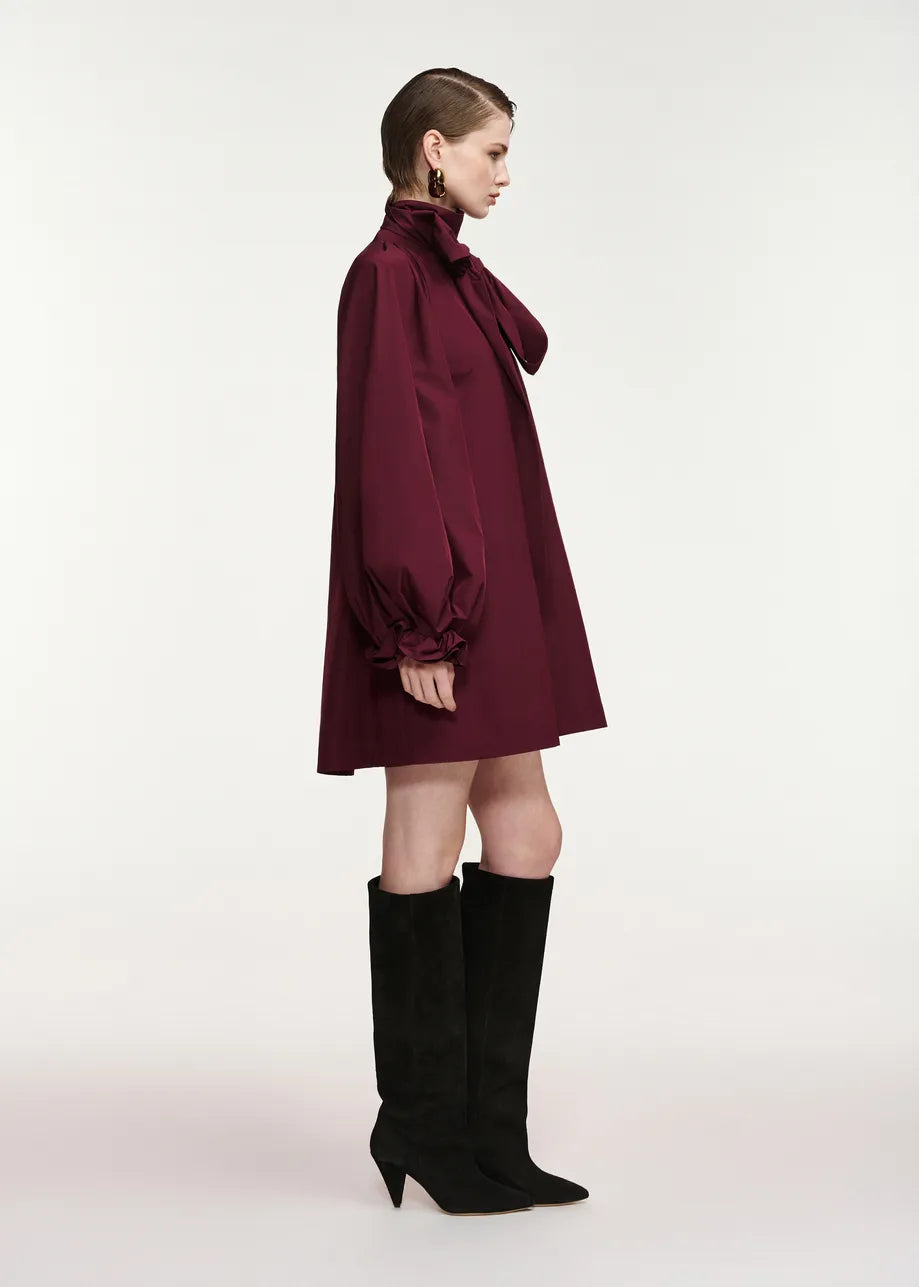 A model is shown from the side wearing a dark burgundy satin-like mini dress with a high collar and voluminous balloon sleeves, paired with black knee-high heeled boots, against a plain light gray studio background.