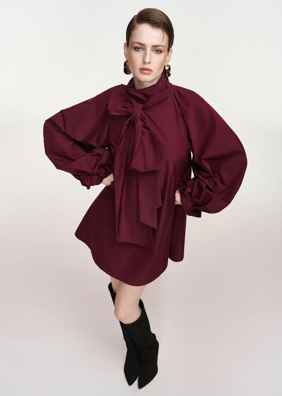 A model wearing a dark burgundy mini dress with dramatic balloon sleeves, a high gathered collar, and a large bow at the neck, paired with black pointed-toe ankle boots against a light gray studio background.