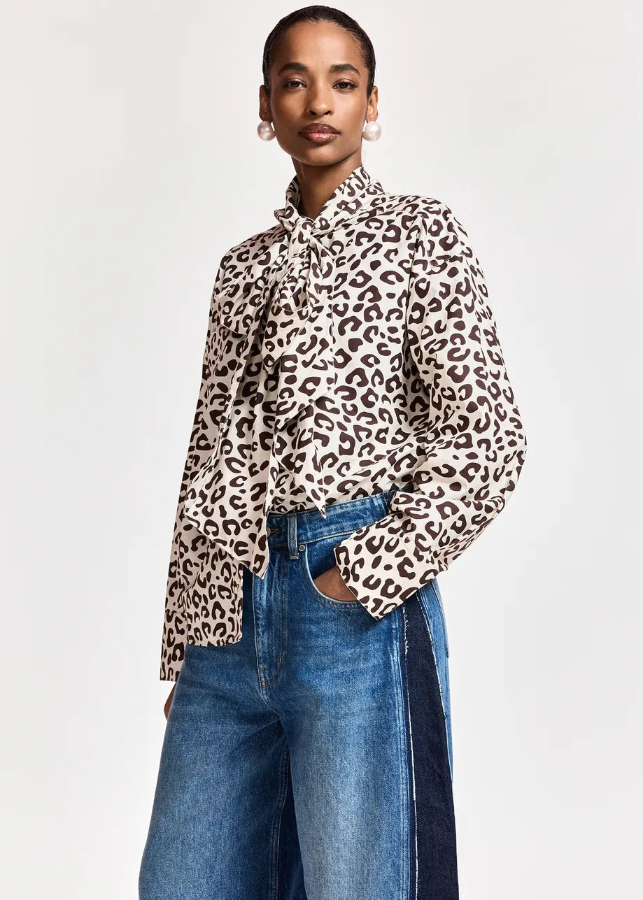 A woman wearing the Essentiel Antwerp ‘Khaleesi’ Leopard Print Pussy Bow Shirt in brown, paired with high-waisted blue jeans.