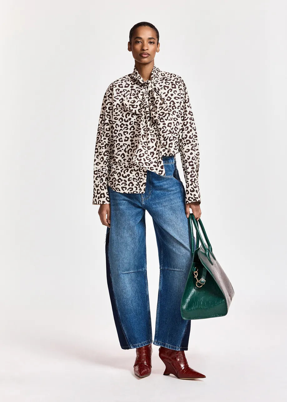 A person stands against a plain background wearing the Essentiel Antwerp Khaleesi Leopard Print Pussy Bow Shirt in brown, paired with wide-leg blue jeans, red ankle boots, and holding a large green handbag.