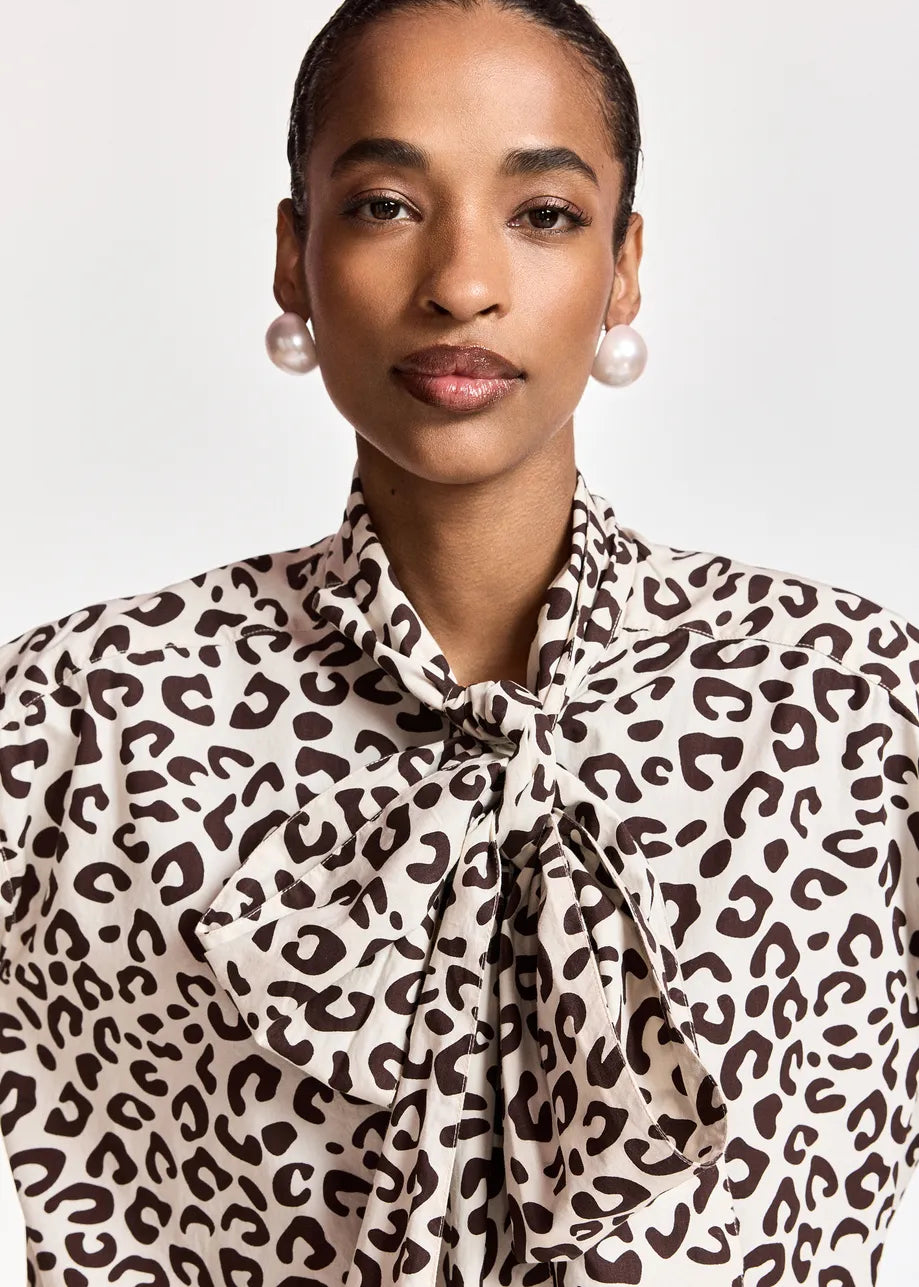 A person wearing the Khaleesi Leopard Print Pussy Bow Shirt in brown by Essentiel Antwerp, styled with pearl earrings and posing against a plain background.