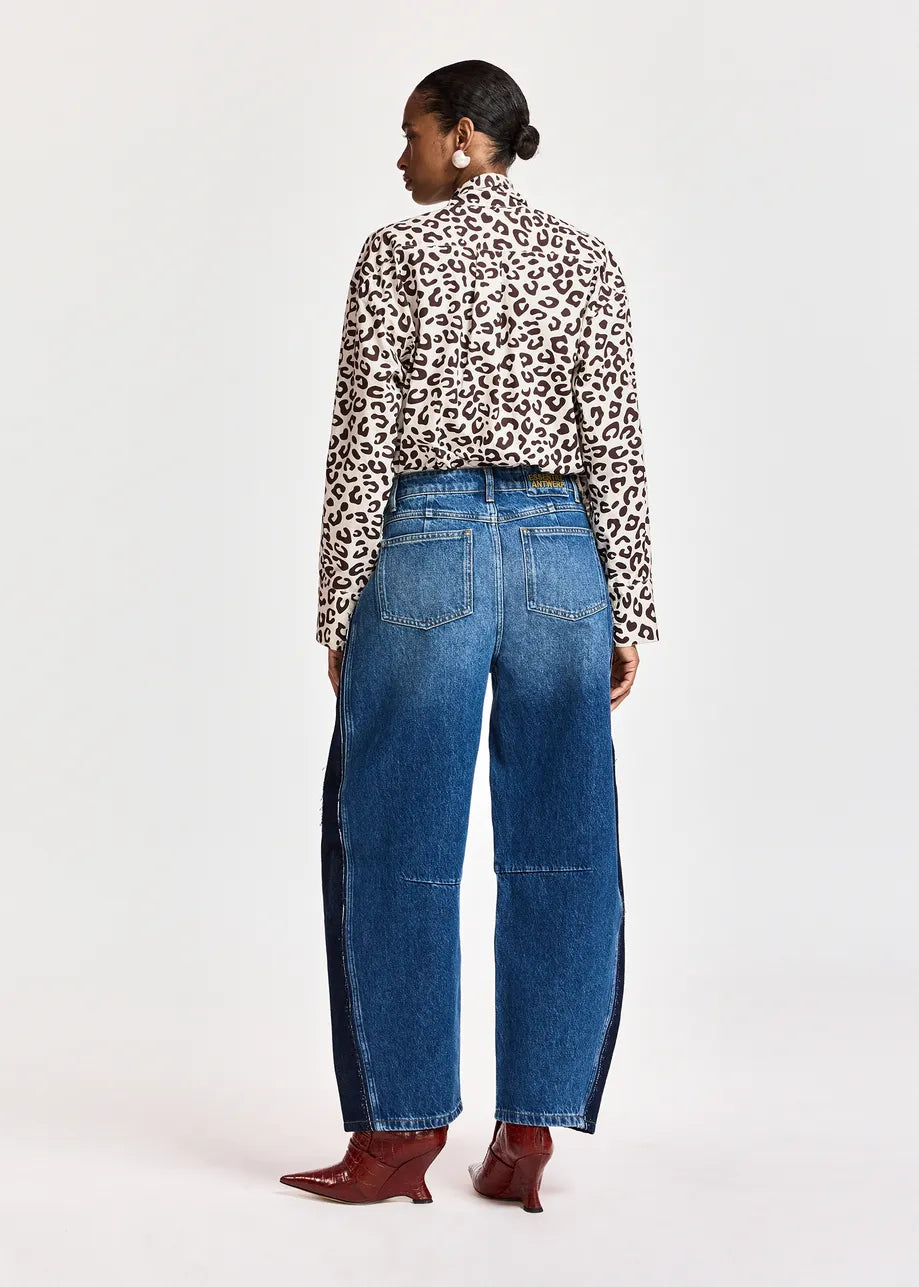 A person stands with their back to the camera against a plain background, wearing the Essentiel Antwerp Khaleesi Leopard Print Pussy Bow Shirt in brown, paired with high-waisted blue jeans featuring dark side panels and pointed brown boots.