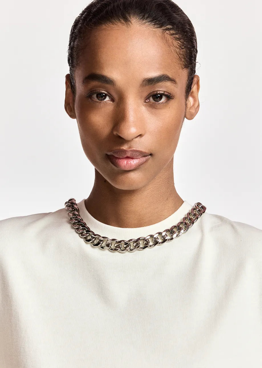A close-up portrait of a model wearing a chunky silver-toned chain necklace over a white crewneck top, set against a plain light gray background.