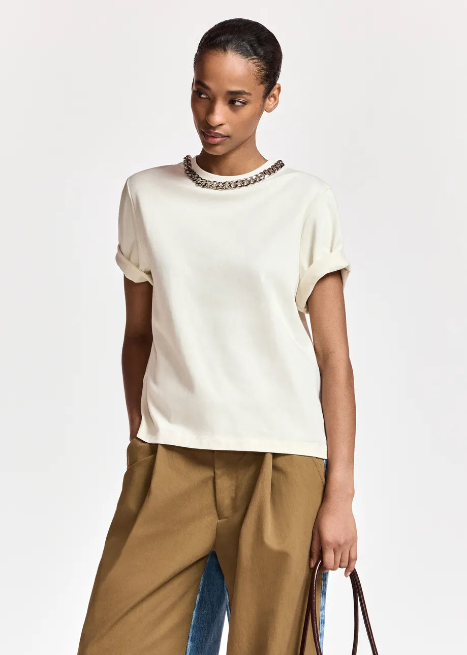 A model wearing a cream-colored short-sleeve knit top with a decorative metallic chain collar, paired with brown trousers, against a plain light gray background.