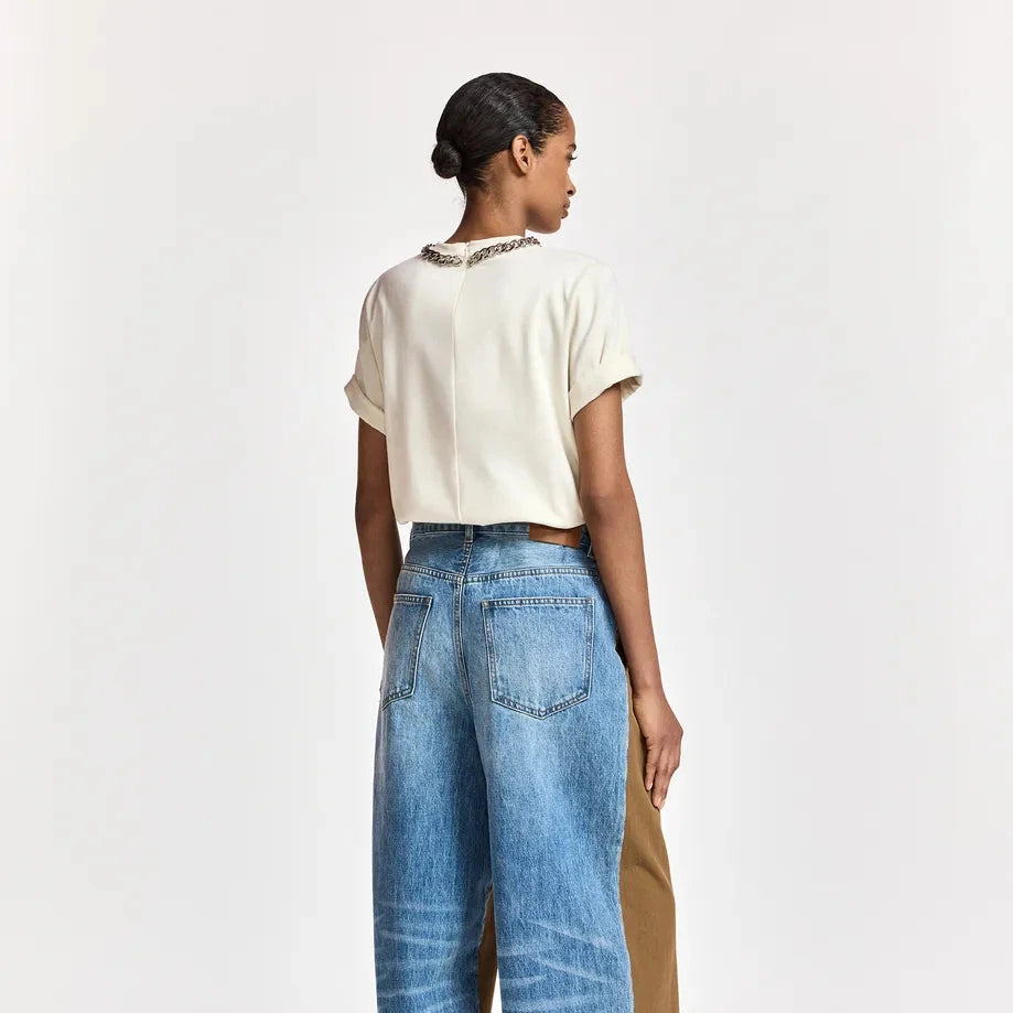 A model shown from the back wearing light-wash wide-leg blue jeans with a brown side panel, a cream short-sleeve top, and red shoes, photographed against a plain light gray studio background.
