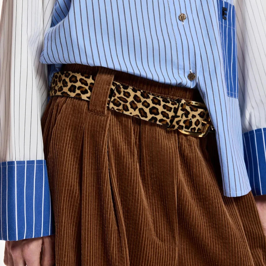 A close-up of a person wearing brown corduroy wide-leg trousers, a blue and white striped button-up shirt, and a leopard-print belt.